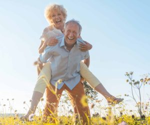 Low-angle view portrait of a happy senior man laughing while carrying his partner on his back, in a sunny day of summer in the countryside