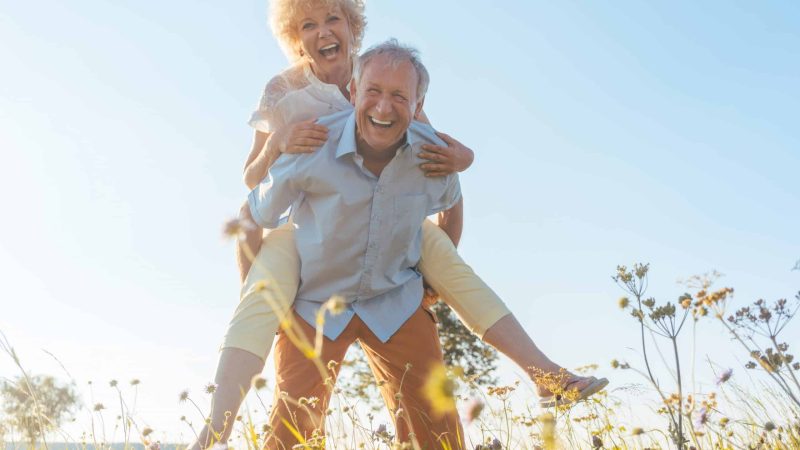 Low-angle view portrait of a happy senior man laughing while carrying his partner on his back, in a sunny day of summer in the countryside